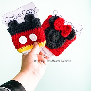 May include: Two crocheted coffee cup cozies, one red and black with a bow and the other red, black, and yellow with two white buttons. The cozies are shaped like Mickey Mouse and Minnie Mouse.