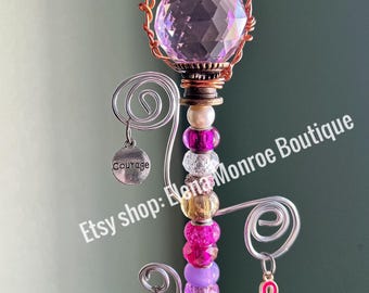 May include: A decorative wand featuring a large faceted crystal ball, surrounded by pink and clear beads. The wand has a copper wire frame and is adorned with various beads in shades of pink, purple, and gold. Charms with the words "Courage" and "Faith" hang from the wand, along with a pink ribbon.