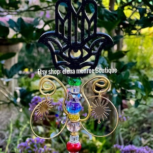 May include: A decorative garden stake featuring a black Hamsa hand design at the top. The stake has a colorful beaded center with gold wire accents and sun-shaped charms. The text "Etsy shop: elena monroe Boutique" is visible.
