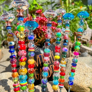 May include: A set of eight colorful garden stakes with crystal tops. Each stake is adorned with a variety of beads, including clear, red, blue, green, yellow, and black beads. The stakes are arranged in a row on a bed of rocks and green plants.