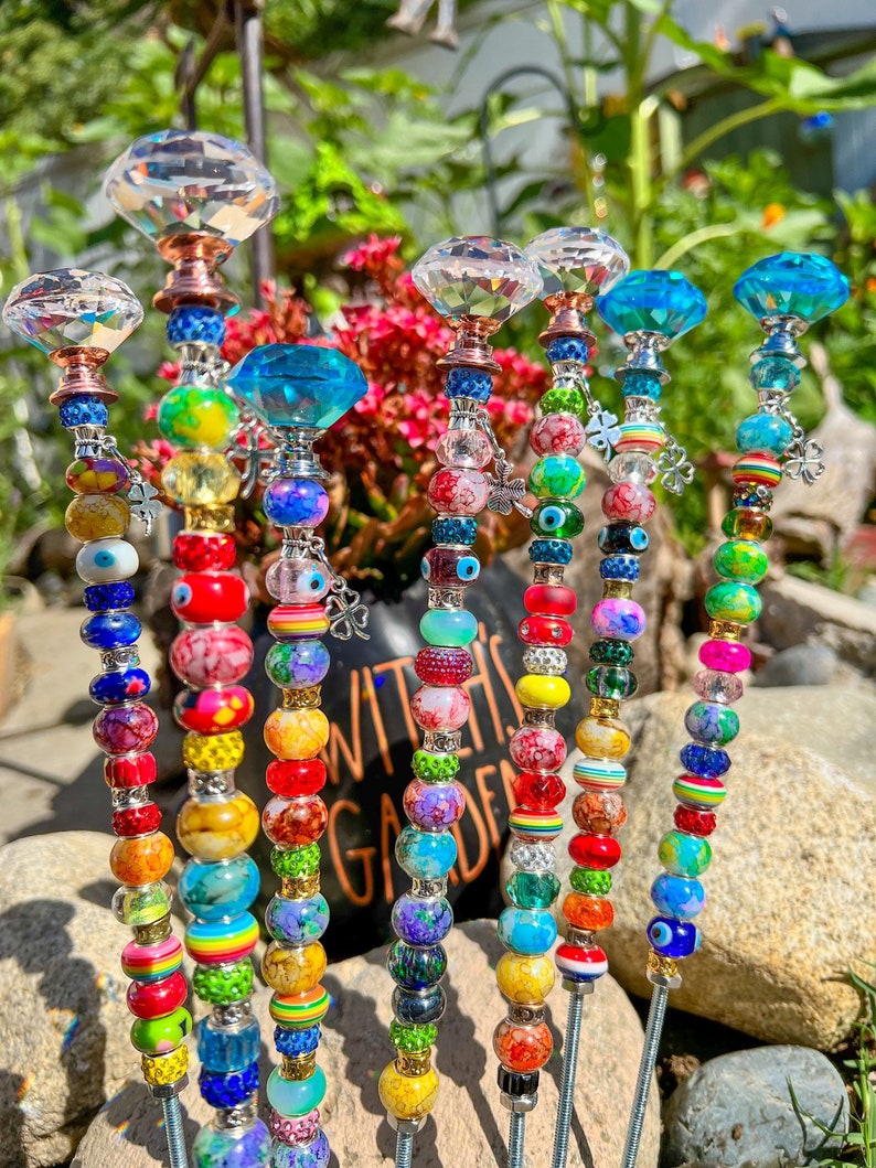 May include: A set of eight colorful garden stakes with crystal tops. Each stake is adorned with a variety of beads, including clear, red, blue, green, yellow, and black beads. The stakes are arranged in a row on a bed of rocks and green plants.