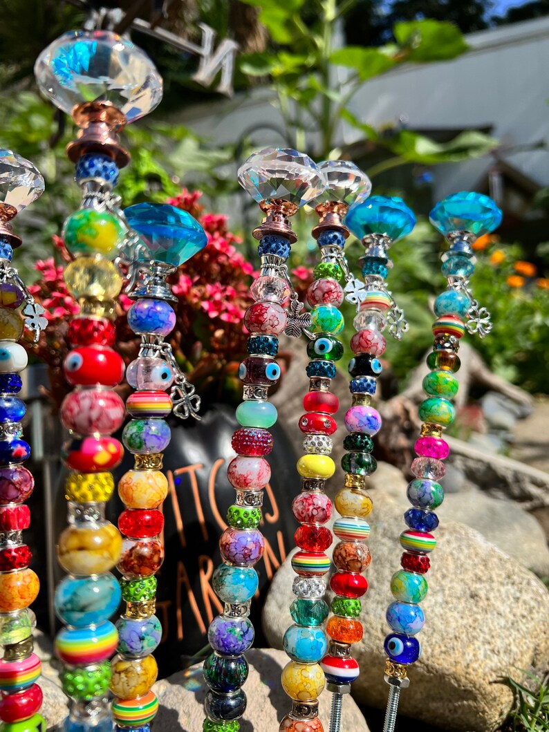 May include: A set of colorful beaded garden stakes with crystal tops. The beads are made of glass and feature various patterns and colors, including red, blue, green, yellow, and white. The stakes are topped with clear crystal shapes.