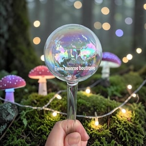May include: A clear glass sphere with an iridescent finish, featuring the text Elena monroe boutique printed in white. The sphere is attached to a slender glass stem, creating a decorative plant watering globe with a reflective surface.