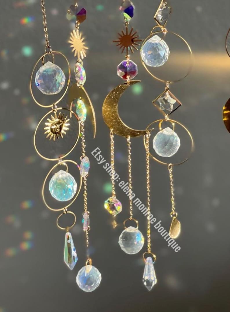 Peut inclure: Attrape-soleil c&eacute;lestes dor&eacute;s avec des accents de cristal. Chaque capteur de soleil pr&eacute;sente un soleil, une lune et des sph&egrave;res de cristal, suspendus par de fines cha&icirc;nes. Le texte "Etsy shop: elena monroe boutique" est visible.