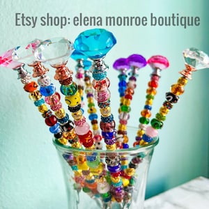 May include: A collection of colorful beaded wands with crystal tops. The wands are arranged in a clear glass vase. The wands are made with various colors of beads, including pink, purple, blue, green, yellow, orange, and red. The crystal tops are clear, blue, and purple.
