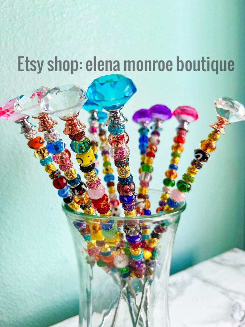 May include: A collection of colorful beaded wands with crystal tops. The wands are arranged in a clear glass vase. The wands are made with various colors of beads, including pink, purple, blue, green, yellow, orange, and red. The crystal tops are clear, blue, and purple.
