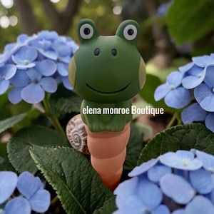 May include: A green frog garden stake with a smiling face and large eyes. The frog is perched atop a terracotta-colored spiral. The text "elena monroe Boutique" is visible. The background features blue hydrangea flowers and green foliage.