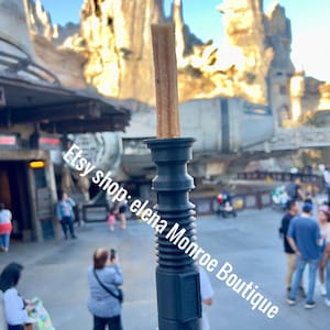 May include: A grey lightsaber-shaped churro holder with a brown churro inside. The holder has a black band around the top and bottom. The background is a blurry image of a Star Wars themed amusement park.