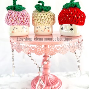 May include: Three ceramic mugs with crocheted strawberry hats on a pink cake stand. The mugs have a white background with a smiling face and closed eyes. The hats are in pink, yellow, and red.