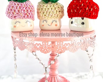 May include: Three ceramic mugs with crocheted strawberry hats on a pink cake stand. The mugs have a white background with a smiling face and closed eyes. The hats are in pink, yellow, and red.