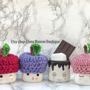 May include: Four white ceramic mugs with smiling faces and crocheted apple hats in red, purple, pink, and a mug with a chocolate drizzle and a marshmallow. The mugs are on a white marble background. Etsy shop: Elena Monroe Boutique