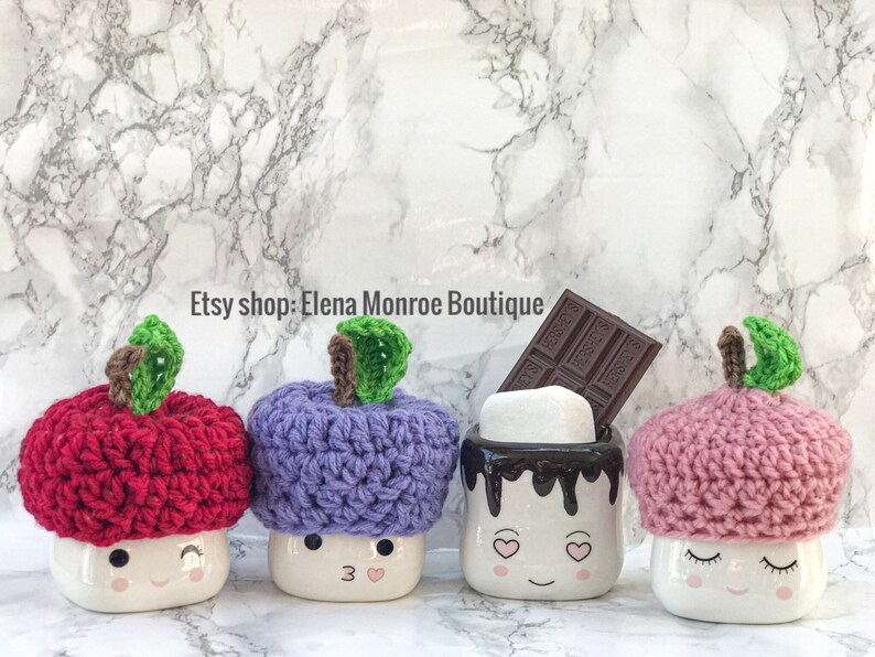May include: Four white ceramic mugs with smiling faces and crocheted apple hats in red, purple, pink, and a mug with a chocolate drizzle and a marshmallow. The mugs are on a white marble background. Etsy shop: Elena Monroe Boutique