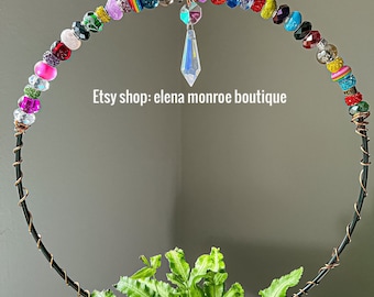 May include: A black metal hoop with colorful beads and a crystal prism hanging from the center. The hoop is decorated with a variety of beads, including clear, red, pink, purple, green, blue, and yellow beads. The hoop is placed over a green plant.
