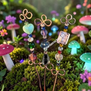 May include: Three decorative garden stakes with copper wire accents and colorful glass beads. Each stake features a unique design, including heart, square, and round beads in shades of green, blue, and purple. The stakes are set against a mossy background with miniature mushrooms.