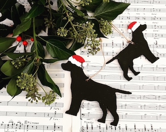 Puede incluir: Dos adornos de perro negro con gorros de Papá Noel rojos y blancos, sobre partituras con follaje verde y bayas rojas. Los adornos están colgados con hilo, creando una decoración festiva para las fiestas.