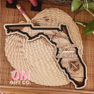 May include: Wooden Florida-shaped wall art with a map design, black border, and text that reads "Hurlburt Field." Includes the text "SRA AFSOC OC A332 SOF PED COORDINATOR JAN 1, 2024 - DEC 31, 2024." The art is placed on a woven fan.