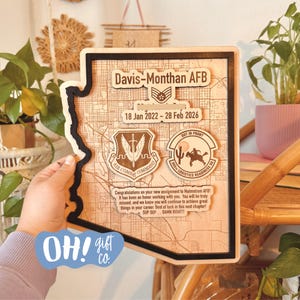 May include: A wooden plaque in the shape of Arizona, with a black border, featuring a map design and military insignia. The plaque includes text: "Davis-Monthan AFB", dates, and a congratulatory message. The lower left corner has a blue sticker with "OH! gift co."