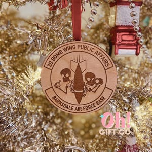 May include: A wooden Christmas ornament with a brown background and a white inscription. The inscription reads "20 Bomb Wing Public Affairs Barksdale Air Force Base". The ornament features a cartoon image of two skeletons holding a bomb with wings.