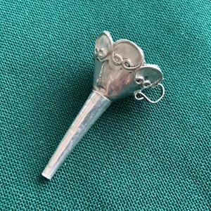 Puede incluir: Un embudo plateado con una parte superior acampanada y un pico largo y cónico. La parte superior presenta formas decorativas en forma de pétalo y un pequeño lazo. El embudo probablemente está hecho de metal y está sobre un fondo texturizado de color verde azulado.