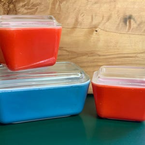 Op de afbeelding: Een set van drie vierkante en rechthoekige glazen bewaarbakjes met doorzichtige glazen deksels. Eén bakje is een grote blauwe rechthoek, terwijl de andere twee kleinere oranje vierkanten zijn. Elk bakje heeft een gladde, effen buitenkant.