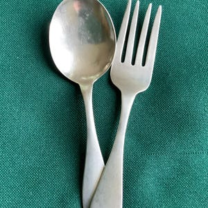 May include: A silver-toned metal spoon and a matching four-tined fork are positioned overlapping each other. Both utensils feature a smooth, plain handle design with a rounded end, showcasing a polished metallic finish that reflects light.