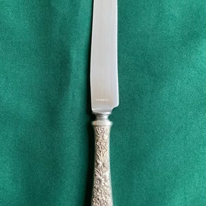 May include: A stainless steel knife with a floral handle design. The silver blade contrasts with the detailed floral pattern on the handle. The knife is displayed against a green backdrop.