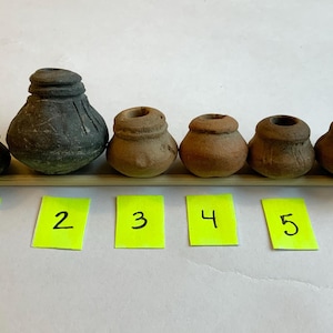 May include: Six small, antique-looking pottery vessels of varying sizes, arranged on a wooden dowel. The vessels are in shades of brown and gray. Each vessel has a rounded body and a small opening at the top. Yellow sticky notes below are numbered 1 through 6.