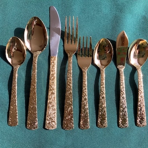 May include: A set of gold-colored flatware, including spoons, forks, and knives, arranged on a green surface. The handles of the utensils have a textured floral pattern. The knife blades are silver.