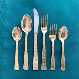 Könnte beinhalten: Ein Set aus sechs goldfarbenen Besteckteilen, darunter Löffel, Gabeln und ein Messer. Die Griffe weisen ein Blumenmuster auf. Das Besteck ist auf einer türkisblauen Oberfläche angeordnet.