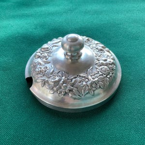 May include: A silver-colored, round lid with a floral design. The lid features a central knob and a detailed relief of flowers and leaves around the edge. The lid has a small notch on the side, and it sits on a green surface.