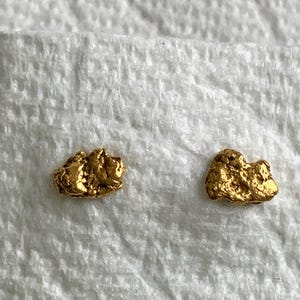 Puede incluir: Dos pequeñas pepitas de oro de forma irregular. Las pepitas tienen una superficie rugosa y texturizada y un color dorado brillante y metálico. Se muestran sobre un fondo blanco texturizado.