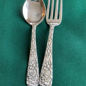 Pode incluir: Um conjunto de colher e garfo de prata com detalhes florais. A colher tem uma tigela arredondada e o garfo tem quatro dentes. Ambos os utensílios têm padrões florais ornamentados nas alças, sobre um fundo verde.