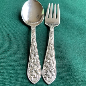 May include: A silver spoon and fork set with floral detailing. The spoon has a rounded bowl, and the fork has three tines. Both utensils have ornate floral patterns on the handles. The set is displayed on a green surface.