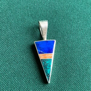 May include: A triangular pendant with a silver bail. The pendant features a mosaic design with blue, orange, and turquoise sections. The pendant is set against a green textured background.