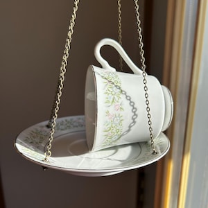 Op de afbeelding: Een witte kop en schotel set met een bloemenpatroon, opgehangen aan een goudkleurige ketting. De kop is gekanteld en rust op de schotel. De set heeft een gouden rand.