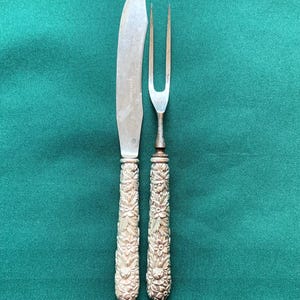 May include: A silver-coloured carving knife and fork set with floral handles. The knife has a curved blade, and the fork has two prongs. The handles are decorated with a detailed floral pattern. The set is presented on a green background.