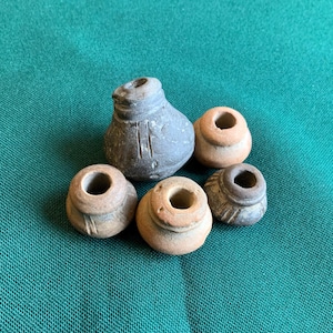 Peut inclure: Un ensemble de cinq petits fusaïoles en argile façonnées à la main. L'une est gris foncé avec des lignes verticales gravées, tandis que les quatre autres sont de couleur beige clair ou terre cuite, avec des formes arrondies et des trous centraux.