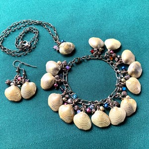 Puede incluir: Un conjunto de joyas que incluye un collar, pendientes y una pulsera. El conjunto incluye pequeñas conchas de color claro unidas a cadenas plateadas. Los pendientes y la pulsera tienen detalles de cuentas de colores. Las joyas se exhiben sobre una superficie verde azulado.