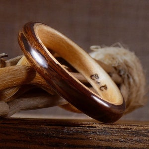 Puede incluir: Un anillo de madera marrón oscuro con una incrustación de madera más clara. El anillo tiene las letras "E & E" grabadas en el interior.