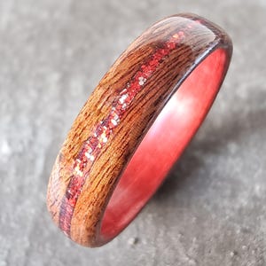 Peut inclure: Une bague en bois avec un intérieur rouge et une incrustation centrale de paillettes rouges. L'extérieur de la bague est brun chaud avec le grain du bois visible. La bague est photographiée en gros plan sur une surface grise.