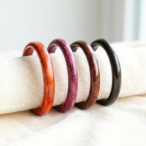 Puede incluir: Cuatro servilleteros de madera en tonos naranja, morado, marrón y negro. Los anillos son lisos y redondos, apoyados sobre una tela blanca. Los colores varían de claro a oscuro, mostrando la veta natural de la madera.