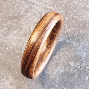 Puede incluir: Un anillo de madera con un patrón de vetas de madera natural en tonos marrón y beige. El anillo tiene un acabado liso y pulido y una forma redondeada. El nombre "Khalida D" está grabado en el interior del anillo.