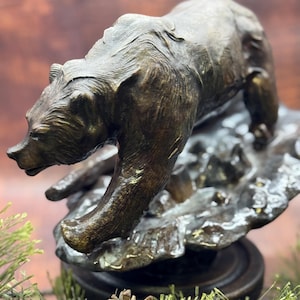 May include: A bronze-toned sculpture of a bear walking across a rocky, uneven terrain. The bear is depicted in a forward-leaning posture with textured fur details, mounted on a circular base. The finish has a weathered, metallic appearance.