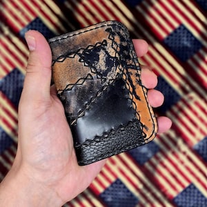 Puede incluir: Una cartera de cuero hecha a mano con un diseño desgastado de dos tonos. La cartera presenta una combinación de cuero negro y marrón claro, cosidos con un hilo en contraste. La cartera se sostiene en una mano.