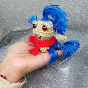 May include: A handmade crochet creature with a tan head, red scarf, and blue hair and tail. The creature has black eyes and is held in a hand. The background is a sparkly silver surface. The text "@Msc_crochet" is visible.