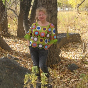 Könnte beinhalten: Eine Frau trägt ein farbenfrohes Häkeltop mit einem kreisförmigen Muster in Grün-, Blau-, Rot- und Weißtönen. Sie steht in einem Waldgebiet mit heruntergefallenen Blättern auf dem Boden.
