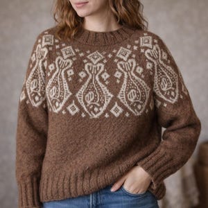 Puede incluir: Un suéter marrón con un patrón geométrico color crema en el pecho y los hombros. El suéter tiene cuello redondo y puños y dobladillo acanalados. El diseño presenta formas estilizadas y patrones de diamantes. El suéter está hecho de un material suave y texturizado.