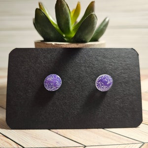 May include: A pair of round stud earrings. Each earring features a purple and white speckled design. The earrings are displayed on a black card, with a small potted succulent in the background.