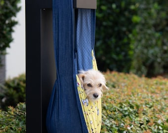 May include: A small dog looks out from a blue denim pet carrier. The carrier is suspended from a black metal post and has a yellow interior with a cartoon pattern. The pet carrier is designed for small dogs.
