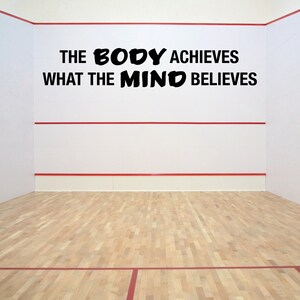 May include: A white wall with a red line across the middle. The wall has a black motivational quote that reads "The body achieves what the mind believes". The wall is in a squash court with a wooden floor.